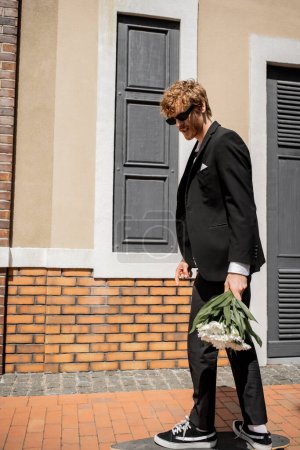 Téléchargez les photos : Élégant homme en costume noir et lunettes de soleil marchant sur la rue de la ville, marié avec bouquet de mariage - en image libre de droit