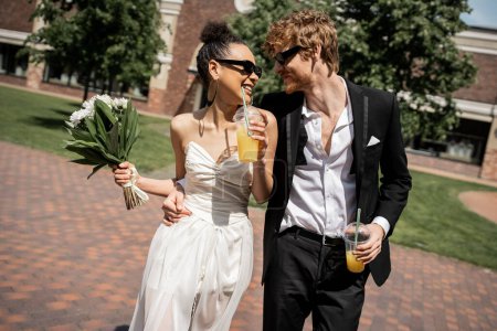 Photo for Joyful multiethnic newlyweds in sunglasses walking with orange juice, wedding on urban street - Royalty Free Image