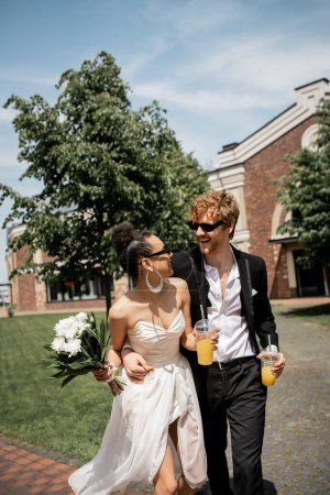 Photo for Diverse couple with orange juice and flowers smiling at each other, wedding on street - Royalty Free Image
