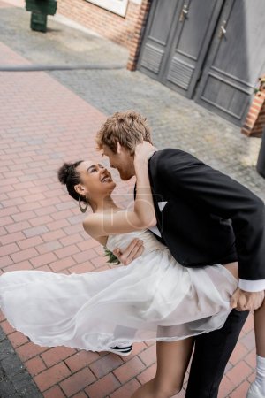 stylish redhead man embracing joyful african american bride, wedding on urban street