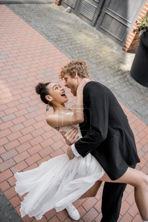 novio elegante joven abrazando excitada novia afroamericana, boda en la ciudad europea