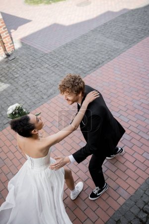 Photo for Happy interracial couple in wedding attire running towards each other on urban street, top view - Royalty Free Image