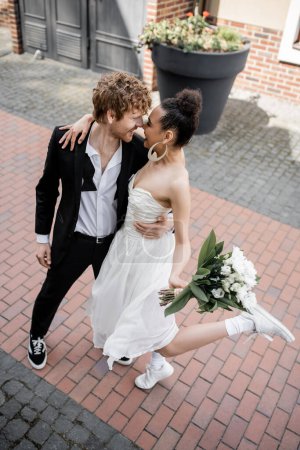 love in city, redhead groom and african american woman with bouquet embracing on street