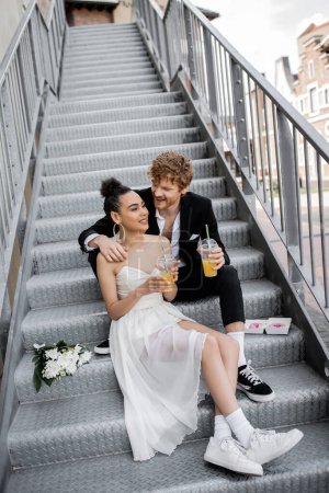Photo for Young and happy interracial couple in wedding attire sitting with orange juice on stairs in city - Royalty Free Image
