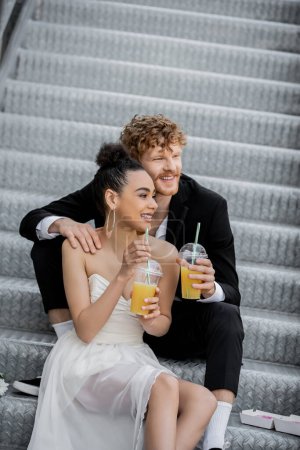 Photo for Redhead groom embracing happy african american bride sitting with orange juice on stairs in city - Royalty Free Image