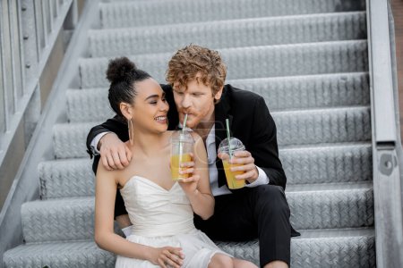 Photo for African american bride smiling near redhead groom drinking orange juice on stairs in city - Royalty Free Image