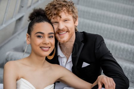 portrait of carefree and stylish interracial newlyweds smiling at camera, outdoor wedding