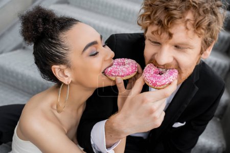 Foto de Jóvenes recién casados multiétnicos se divierten y se alimentan mutuamente con rosquillas dulces en la calle urbana - Imagen libre de derechos