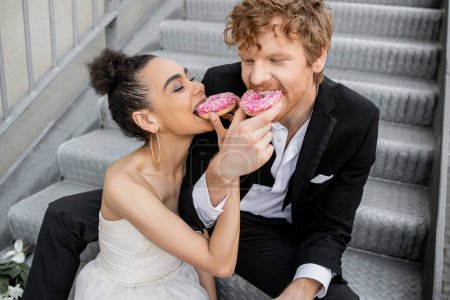 Foto de Celebración de la boda en la ciudad, pareja interracial alimentándose mutuamente con rosquillas dulces en las escaleras - Imagen libre de derechos