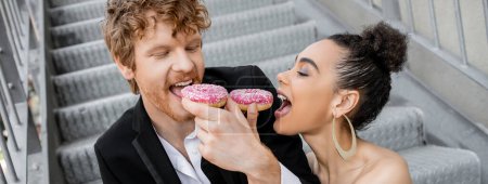 Foto de Boda, entorno urbano, diversión, pareja recién casada interracial alimentándose con rosquillas, pancarta - Imagen libre de derechos