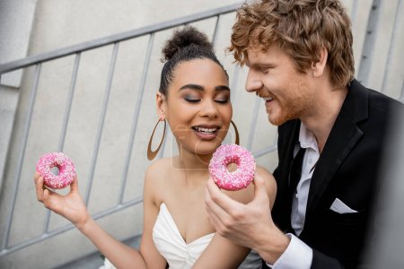 delighted multiethnic couple in wedding outfit holding sweet donuts, wedding in urban setting
