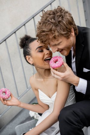 Photo for Cheerful multiethnic newlyweds biting sweet donut, wedding celebration on city street - Royalty Free Image