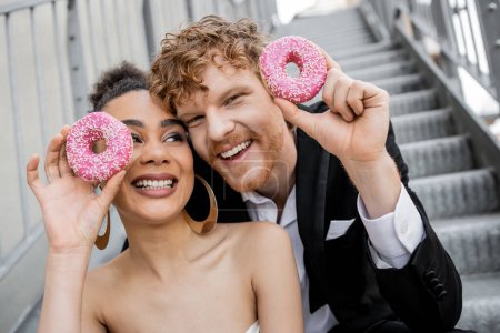 entzückte und elegante multiethnische Brautpaare posieren mit süßen Donuts, Hochzeit in urbaner Umgebung