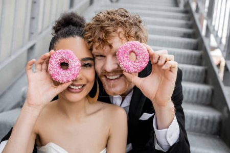 Photo for Having fun, wedding in city, excited interracial newlyweds obscuring face with donuts - Royalty Free Image