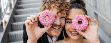 Photo for Outdoor wedding, happy interracial couple obscuring face with sweet donuts, banner - Royalty Free Image
