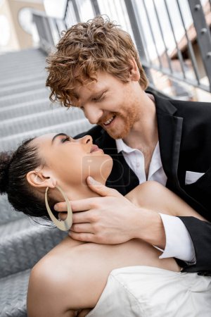 Téléchargez les photos : Romance urbaine, mariage, tendresse, marié rousse heureux embrassant élégante mariée afro-américaine - en image libre de droit