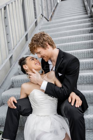 elegant redhead man embracing delighted african american bride on stairs in city