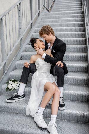urban romance, love, outdoor wedding, young interracial couple embracing near flowers on stairs