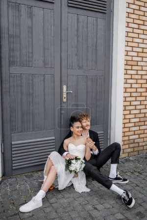 happy interracial couple sitting on street pavement near doors, wedding attire, flowers