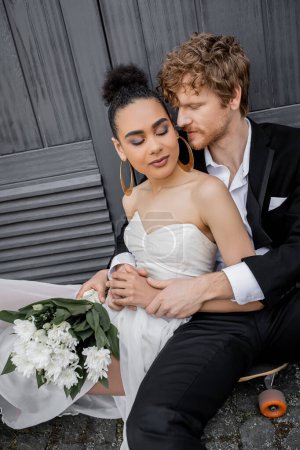 Photo for Redhead groom hugging african american bride with closed eyes and flowers, sitting near building - Royalty Free Image