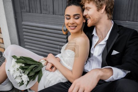 happy young man embracing elegant african american bride sitting with flowers near city building