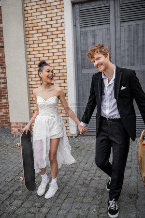 fröhliche gemischtrassige Brautpaare, die mit Longboard und Skateboard auf der Stadtstraße spazieren gehen, Hochzeitskleid