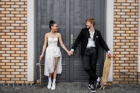 jeunes mariés multiethniques insouciants avec longboard et skateboard tenant la main près de la construction de la ville