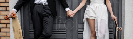 Téléchargez les photos : Vue recadrée du marié avec longboard tenant la main de la mariée afro-américaine près du bâtiment, bannière - en image libre de droit