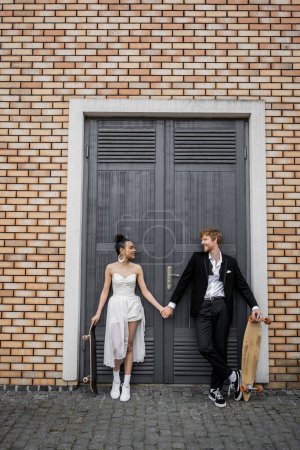 Téléchargez les photos : Joyeux interracial jeunes mariés avec longboard et skateboard regardant les uns les autres près du bâtiment de la ville - en image libre de droit