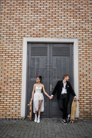 despreocupados recién casados multiétnicos con longboard y monopatín tomados de la mano cerca del edificio de la ciudad