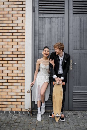 Téléchargez les photos : Jeune couple interracial avec planche à roulettes et longboard près de la construction de la ville, bonheur, rire - en image libre de droit