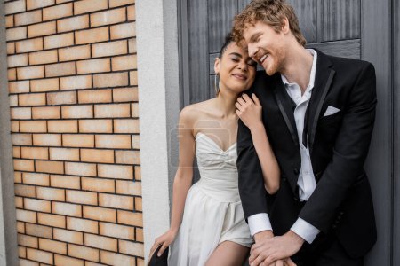 Téléchargez les photos : Enchantée mariée afro-américaine embrassant marié rousse heureux près du bâtiment de la ville, les yeux fermés - en image libre de droit