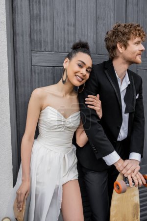 Téléchargez les photos : Couple interracial avec longboard et skateboard en ville, afro-américaine mariée souriant à la caméra - en image libre de droit