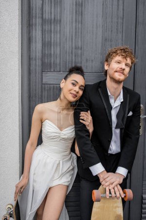 Téléchargez les photos : Afro-américaine mariée souriant à la caméra près de marié rousse près de bâtiment, longboard, planche à roulettes - en image libre de droit