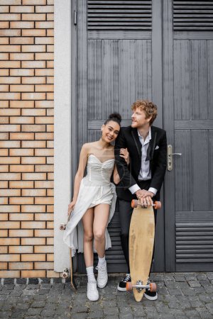 Téléchargez les photos : Couple interracial en tenue de mariage, avec longboard et skateboard près des portes du bâtiment de la ville - en image libre de droit