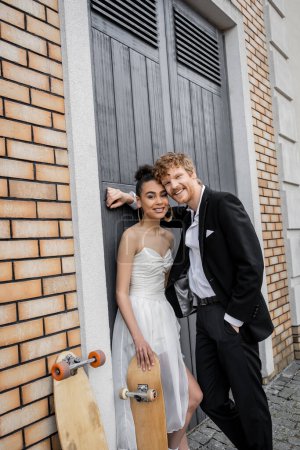 happy redhead groom with hand in pocket, african american bride with skateboard smiling at camera