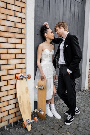 Téléchargez les photos : Élégant marié rousse avec la main dans la poche près de mariée afro-américaine, longboard et skateboard - en image libre de droit