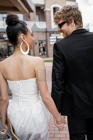 marié rousse en lunettes de soleil, mariée afro-américaine avec longboard se regardant dans la rue