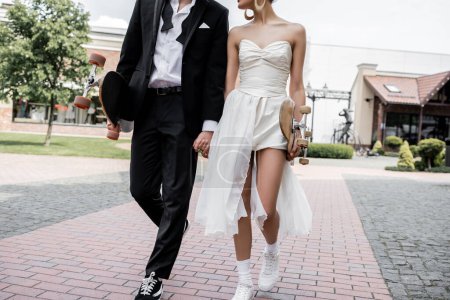 Téléchargez les photos : Vue recadrée d'un élégant couple multiethnique marchant avec longboard et skateboard, mariage en ville - en image libre de droit