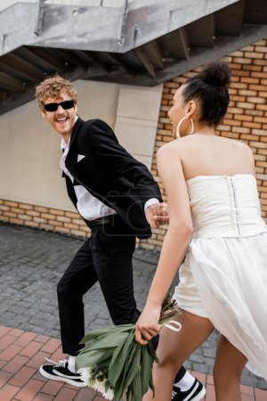 excitado novio en gafas de sol de la mano de la novia afroamericana y corriendo en la calle urbana
