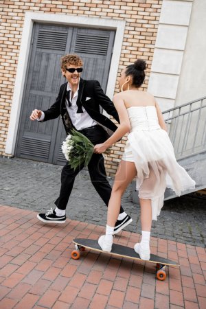 excited groom in sunglasses near african american bride in wedding dress riding longboard