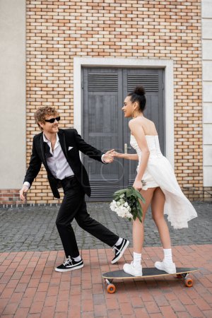 Photo for Redhead groom in sunglasses holding hands with african american bride with bouquet riding longboard - Royalty Free Image