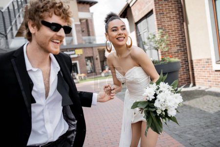 Téléchargez les photos : Afro-américaine mariée américaine avec des fleurs et marié rousse dans des lunettes de soleil tenant la main sur la rue - en image libre de droit