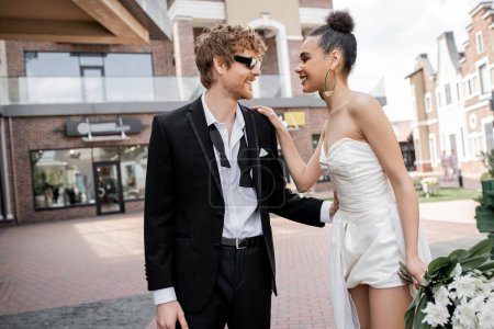 Photo for Outdoor wedding, modern and stylish interracial newlyweds looking at each other on street - Royalty Free Image
