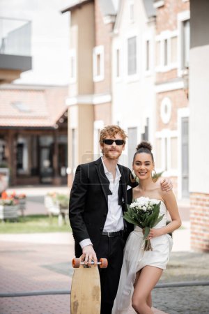 Téléchargez les photos : Mariage en plein air, ville européenne, jeunes mariés interracial élégant avec longboard et bouquet dans la rue - en image libre de droit