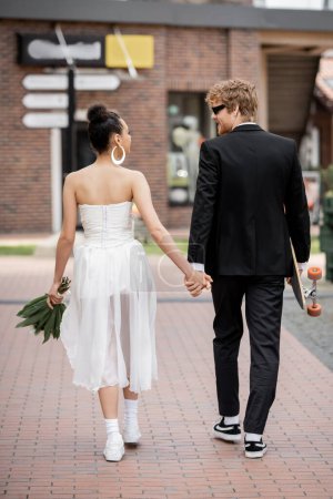 Téléchargez les photos : Vue arrière du couple multiethnique avec longboard et fleurs tenant la main, marchant en ville, mariage - en image libre de droit