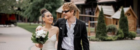 Téléchargez les photos : Mariage en plein air, mariée afro-américaine avec des fleurs et marié dans des lunettes de soleil marchant en ville, bannière - en image libre de droit
