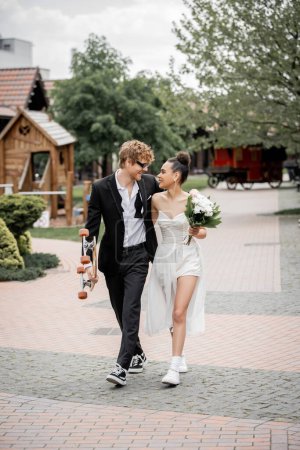 Photo for Happy and stylish multiethnic couple walking with longboard and flowers, smiling at each other - Royalty Free Image