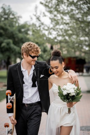 Photo for Redhead groom in sunglasses embracing african american bride, walking with longboard and flowers - Royalty Free Image
