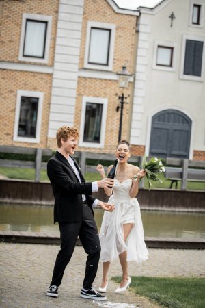 Téléchargez les photos : Marié avec champagne coulant de bouteille près de la mariée afro-américaine ravie, célébration en plein air - en image libre de droit